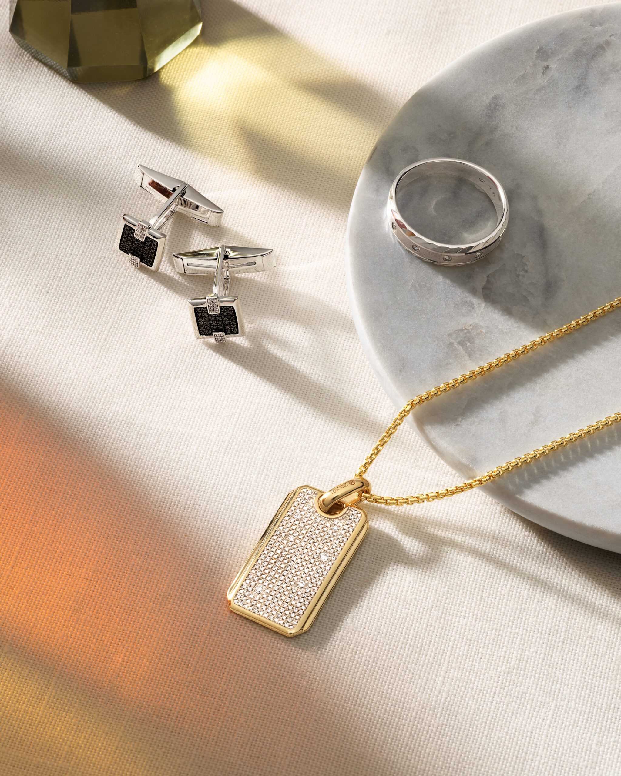 A selection of jewelry for men, including cufflinks, a silver band, and a gold dogtag pendant on a chain.